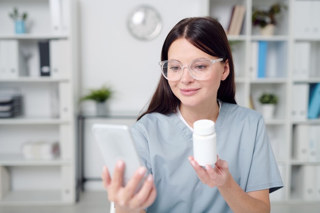 Junge Ärztin in Uniform und Brille, die Smartphone hält und dem Online-Patienten eine Plastikflasche mit neuen wirksamen Medikamenten zeigt