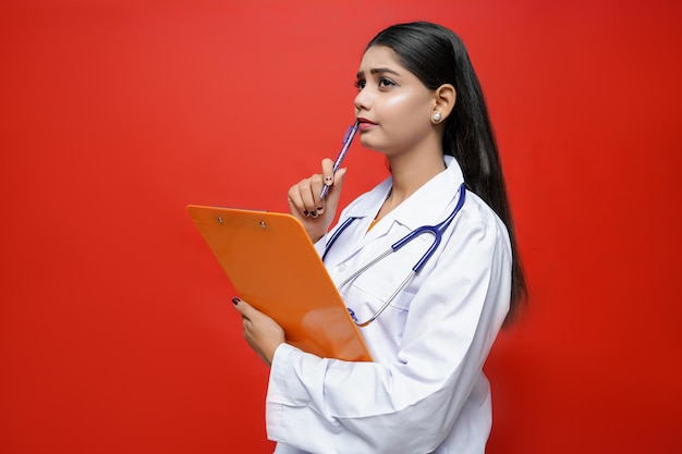 junge Ärztin mit Klemmbrett denken auf rotem Hintergrund indisches pakistanisches Modell