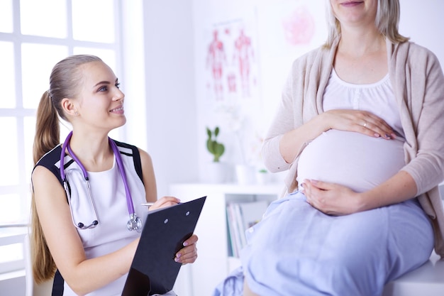 Junge Ärztin mit Stethoskop und Tablet im Gespräch mit schwangerer Frau im Krankenhaus