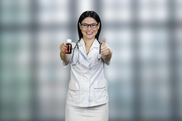 Junge Ärztin zeigt Medizinflasche und gibt Daumen nach oben, Werbung und Werbung verschwommen c