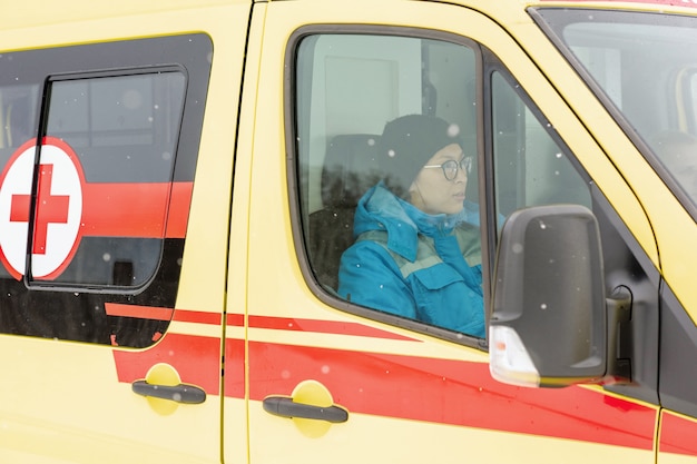 Junge Sanitäterin in Uniform und Mütze sitzt im Krankenwagen, während sie sich beeilt, um zu helfen und kranke Person zu retten