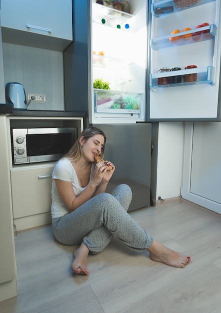 Junge schlaflose Frau sitzt auf Küchenboden neben offenem Kühlschrank und isst Pizza