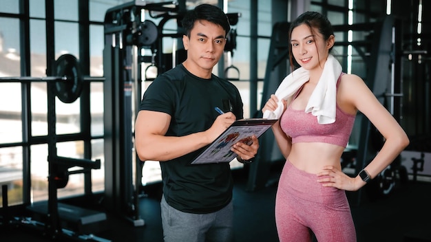 Junge schlanke Fitness-Asiatin, die mit einem Handtuch steht, schaut in die Kamera in der Nähe eines gutaussehenden Trainers, während sie ihren Zeitplan im Fitnessstudio zeigt Der Athlet führt ein gesundes Lebensstil-Cardio-Training zur Gewichtsabnahme durch