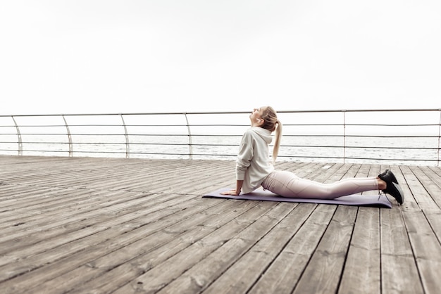 Junge, schlanke Frau, die Yoga-Asana-Pose am Strand praktiziert. Eignungsmädchen, das Übung auf Matte tut. Kobra-Pose