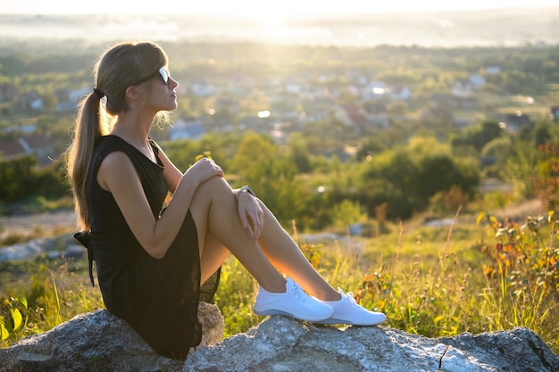 Junge schlanke Frau im schwarzen kurzen Kleid sitzt auf einem Felsen, der sich bei Sommersonnenuntergang im Freien entspannt.