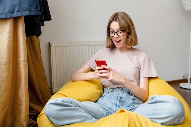 Junge schockierte Frau, die auf Handy in der Wohnung starrt, die rosa Jeans rosa T-Shirt und Brille trägt