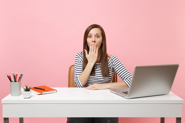 Junge schockierte frau in freizeitkleidung, die den mund mit palmen bedeckt, sitzt am weißen schreibtisch mit zeitgenössischem pc-laptop einzeln auf pastellrosa hintergrund. erfolgsgeschäftskarrierekonzept. platz kopieren.