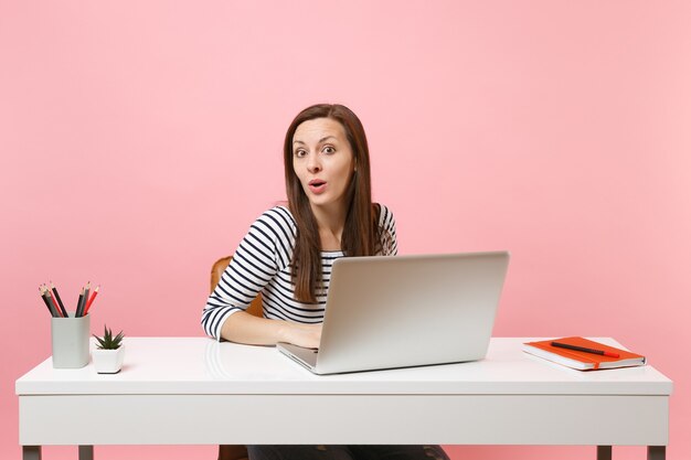 Junge schockierte Frau in Freizeitkleidung, die mit Laptop an einem Projekt arbeitet, während sie im Büro sitzt, isoliert auf pastellrosa Hintergrund. Erfolgsgeschäftskarrierekonzept. Kopieren Sie Platz für Werbung.