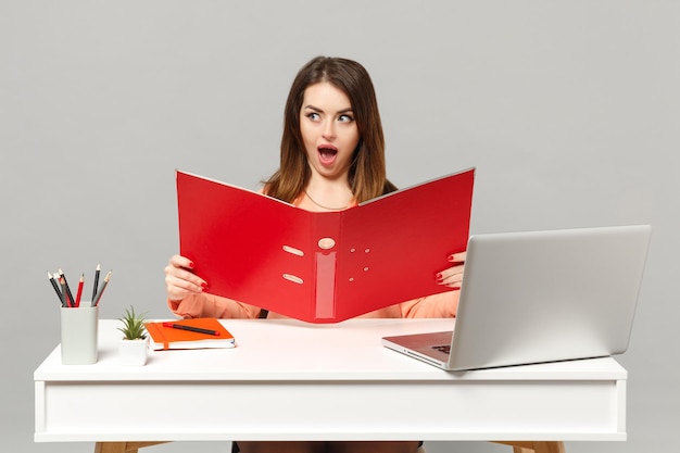 Junge schockierte Frau in pastellfarbener Kleidung mit rotem Ordner für Papiere dokumentiert die Arbeit am Schreibtisch mit PC-Laptop isoliert auf grauem Hintergrund. Leistung Business-Karriere-Lifestyle-Konzept. Mock-up-Kopienbereich.