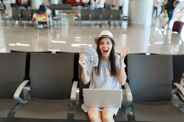 Junge schockierte Reisende Touristenfrau, die am Laptop arbeitet, hält ein Bündel Dollar, Bargeld verteilte Hände warten in der Lobbyhalle am Flughafen?