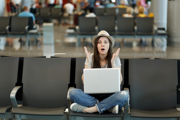 Junge schockierte Reisende Touristenfrau mit Laptop sitzt mit gekreuzten Beinen und spreizt die Hände, während sie in der Lobbyhalle am Flughafen wartet