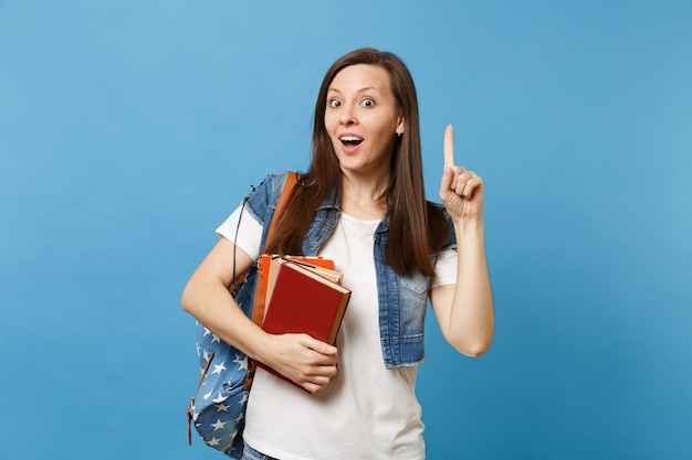 Junge schockierte Studentin mit Rucksack, die mit dem Zeigefinger auf Kopienraum zeigt und eine neue Idee hat, Schulbücher einzeln auf blauem Hintergrund zu halten. Bildung im Hochschulkonzept der High School.