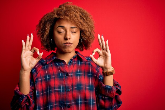 Junge schöne afroamerikanische Afro-Frau mit lockigem Haar, lässigem Hemd, entspannt sich und lächelt mit geschlossenen Augen und macht Meditationsgeste mit den Fingern Yoga-Konzept