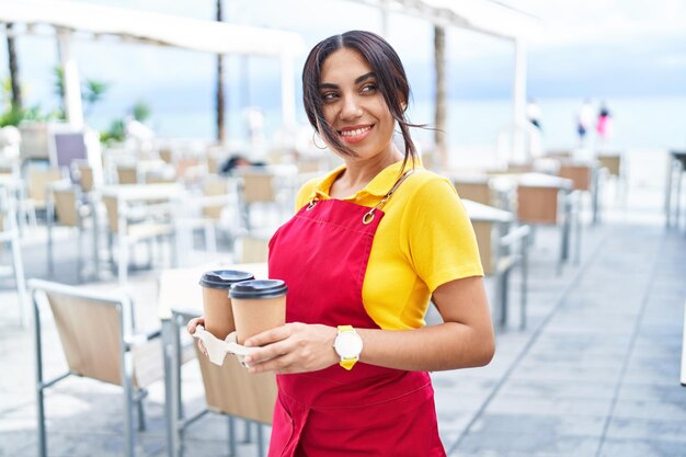 Junge schöne arabische Kellnerin lächelt selbstbewusst und hält Kaffee zum Mitnehmen auf der Terrasse des Cafés