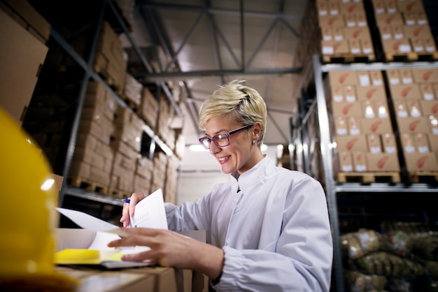 Junge schöne Arbeiterin betrachtet ihre Papiere und lächelt im Fabriklagerraum.