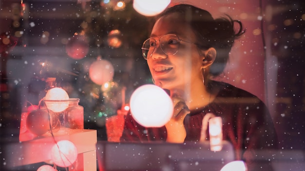 Junge schöne Asiatin fühlen sich mit den Neujahrsfeiern mit Geschenkbox im Haus gut, mit Weihnachtsbaum zu verzieren Konzept frohe Feiertage Glasfensterreflexion und Schneeeffekt.