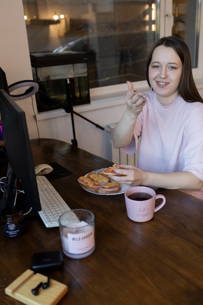 Junge schöne asiatische Bloggerin, die zu Hause am Computer arbeitet und Sandwiches isst und Kaffee trinkt