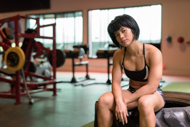 Junge schöne asiatische Fitnessfrau, die im Fitnessstudio sitzt