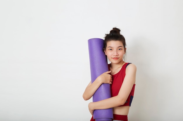 Junge schöne asiatische Frau, die lila Yogamatte steht und hält und bereit ist, im Fitnessstudio zu trainieren, stilvolle Sportbekleidung tragend.