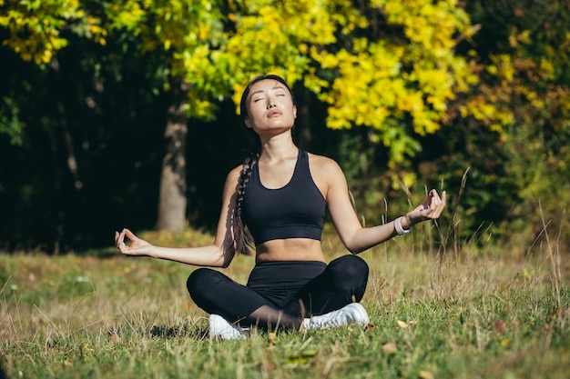 Junge schöne asiatische Frau führt Fitness-Yoga-Übungen durch, sitzt in Lotusposition auf dem Gras und meditiert im Park