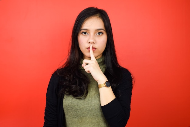 Junge schöne asiatische Frauen, die einen schwarzen Pullover tragen, zeigen ein Schweigezeichen mit rotem, isoliertem Hintergrund