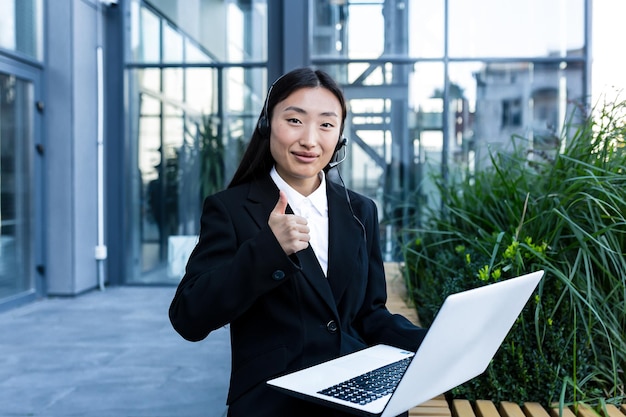 Junge schöne asiatische Geschäftsfrau, die an einem Laptop in der Luft arbeitet, verwendet ein Headset für einen Videoanruf und sitzt in der Nähe eines modernen Büros