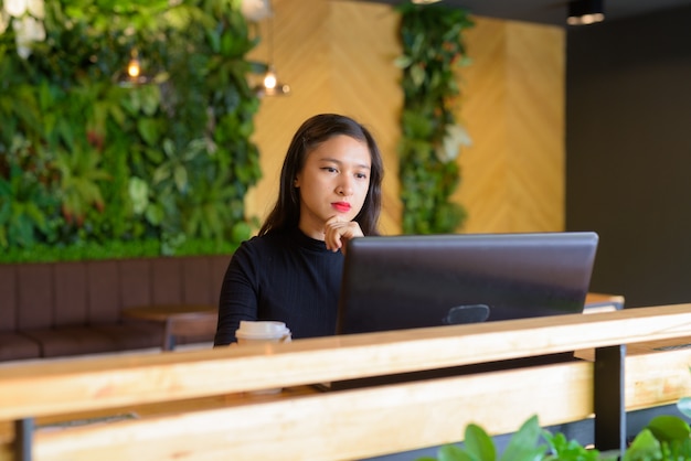 Junge schöne asiatische Geschäftsfrau, die beim Verwenden des Laptops am Kaffeehaus denkt