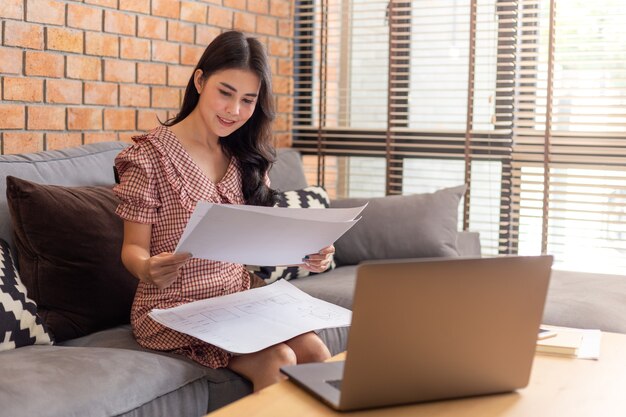 Junge schöne asiatische Geschäftsfrau, die ihre Arbeitspläne vor ihrem Laptop in ihrem Wohnzimmer betrachtet, während sie von zu Hause aus während der COVID-Sperre arbeitet