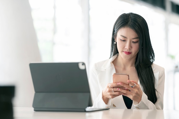 Junge schöne asiatische Geschäftsfrau, die Smartphone beim Sitzen an ihrem Schreibtisch verwendet.