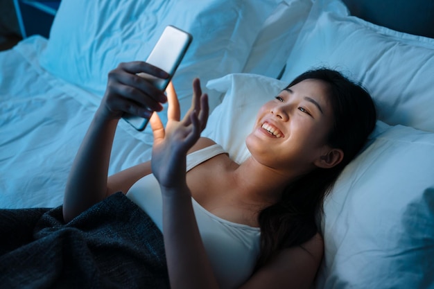 junge schöne asiatische japanische Frau im Bett mit Handy spät in der Nacht dunkles Schlafzimmer. glückliches Mädchen, das im Schlaf liegt, entspannen Sie sich und genießen Sie das Social Media-Netzwerk auf dem Smartphone. Internet-Sucht-Konzept