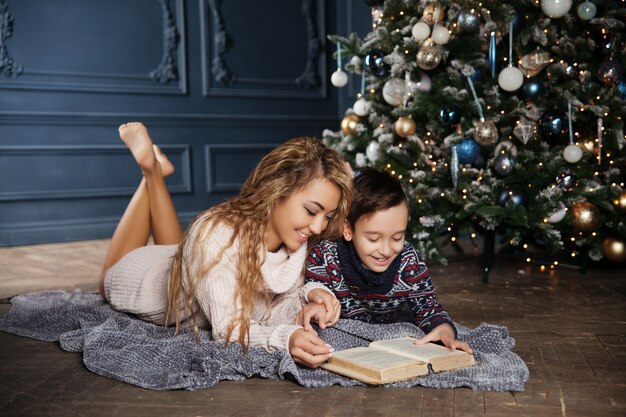 Junge schöne asiatische Mutter mit ihrem kleinen Sohn, der nahe einem verzierten Weihnachtsbaum sitzt und ein Buch liest.