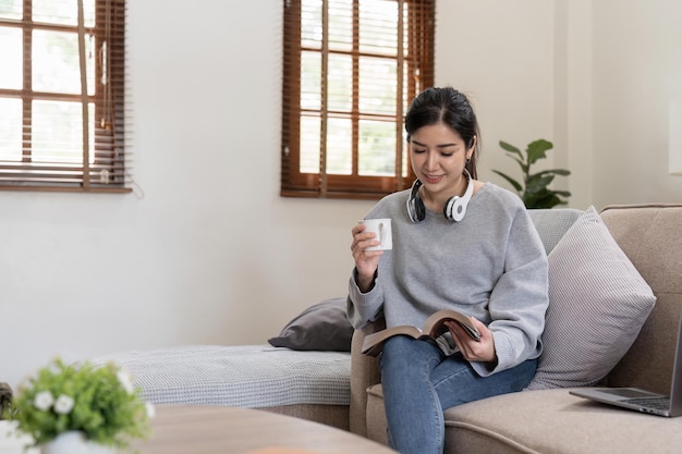 Junge schöne asiatische Studentin sitzt auf dem Sofa und liest zu Hause ein Buch, während sie ihren Laptop zum Online-Lernen nutzt