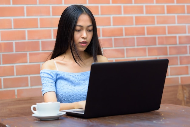 Junge schöne asiatische Teenagerfrau, die Laptop mit Kaffeetasse auf Holztisch gegen Backsteinmauer verwendet