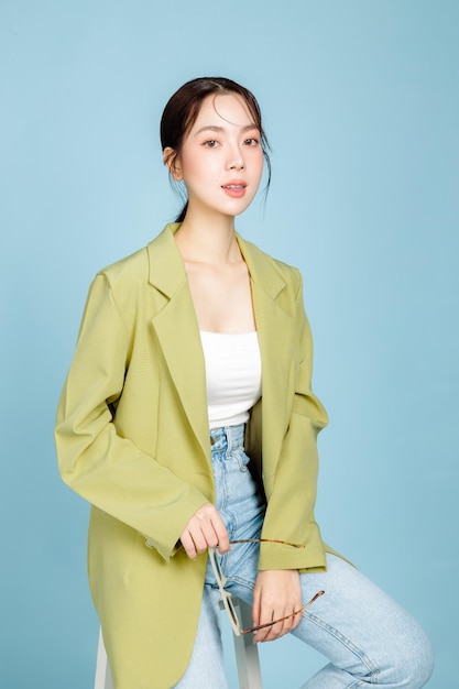 Junge schöne attraktive asiatische Frau in lässiger grüner Modejacke, die auf einem Stuhl sitzt, der auf pastellblauem Hintergrund isoliert ist Weibliches Konsumismus-Lifestyle-Konzept