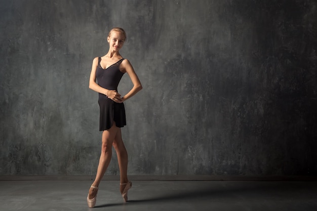 Junge schöne Ballerina in einem schwarzen Kleid, weißen Strumpfhosen und Spitzenschuhen, die wunderschön in einem Bild in einer dunklen Tanzbühne posieren