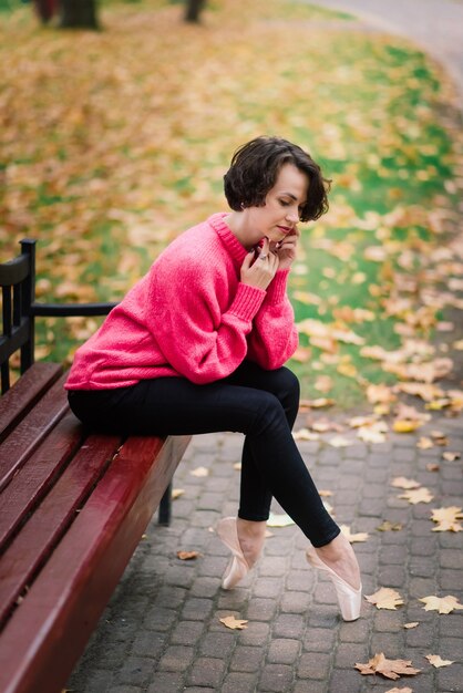 Junge schöne Ballerina in hellem Mantel und Spitzenschuhen sitzen auf der Bank, ruhen draußen im Herbstpark.