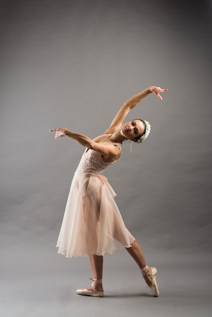 Junge schöne Balletttänzerin im beigen Badeanzug, die auf Punkten auf hellgrauem Studiohintergrund aufwirft
