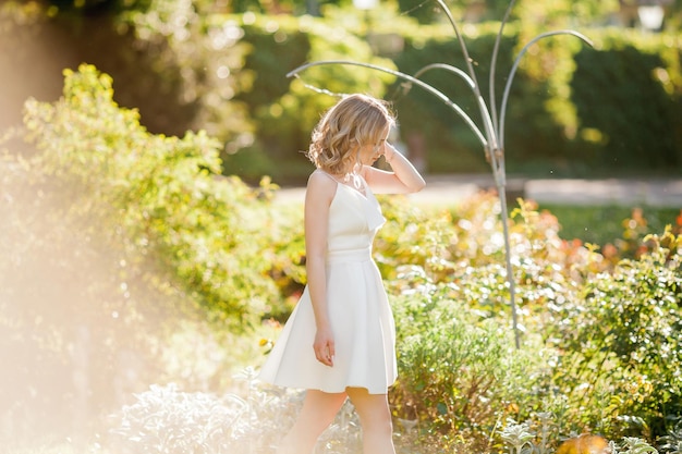 Junge schöne blonde 20er mit lockigem Haar in einem weißen Kleid in den sonnigen Sonnenuntergangsstrahlen im Park Ein stylisches Model geht im Sommer durch den blühenden Park der Stadt