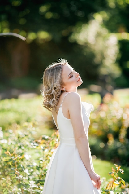 Junge schöne blonde 20er mit lockigem Haar in einem weißen Kleid in den sonnigen Sonnenuntergangsstrahlen im Park Ein stylisches Model geht im Sommer durch den blühenden Park der Stadt