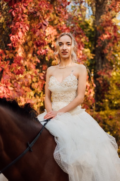 Junge schöne blonde Braut in einem weißen prächtigen Kleid, das ein Pferd im Herbstwald reitet