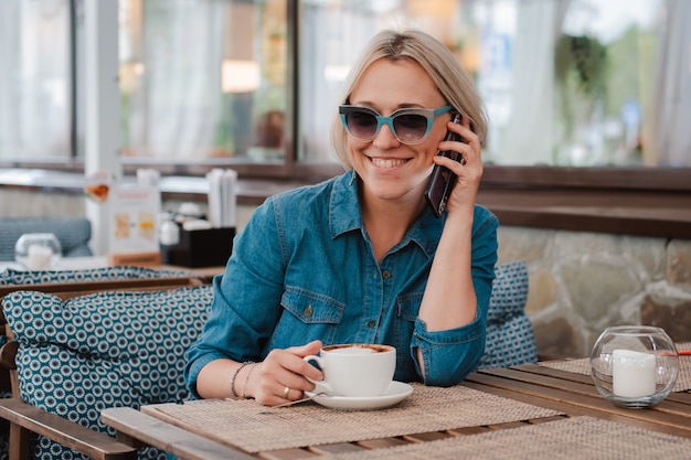 Junge schöne blonde Frau an einem Sommermorgen in der Sonnenbrille kommuniziert auf einem Mobiltelefon in einem Café über einer Tasse Cappuccino-Kaffee. .