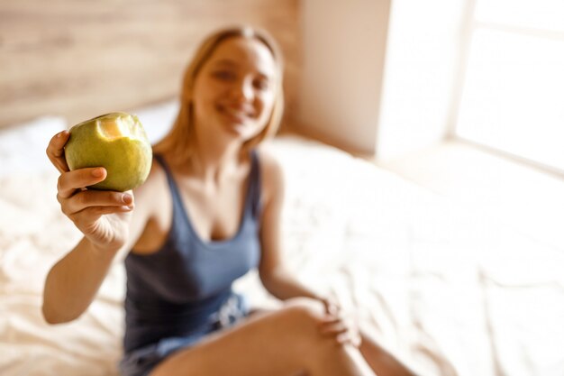 Junge schöne blonde Frau, die am Morgen im Bett sitzt. Unscharfer Hintergrund. Modell halten gebissenen Apfel und zeigen es der Kamera. Fröhlich positiv. Tageslicht.