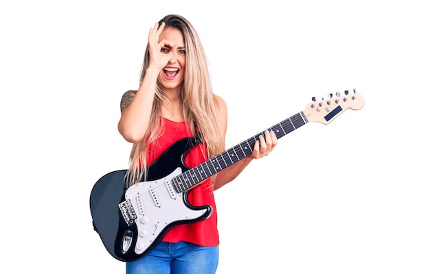 Junge schöne blonde Frau, die E-Gitarre spielt, lächelt glücklich und macht ein Ok-Zeichen mit der Hand auf dem Auge, das durch die Finger schaut