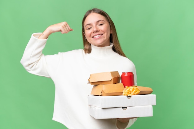 Junge schöne blonde Frau, die Essen zum Mitnehmen vor isoliertem Hintergrund hält und eine starke Geste macht