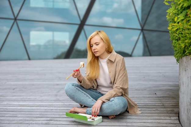 Junge schöne blonde Frau, die Sushi im Freien auf der Holzterrasse neben einem modernen Gebäude in der Stadt isst. Leckeres Essen zum Mitnehmen