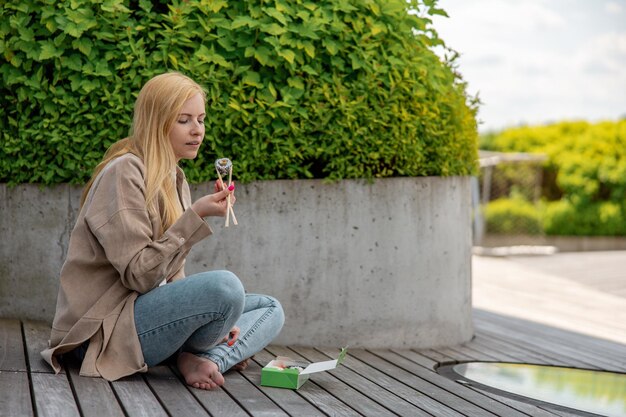 Junge schöne blonde Frau, die Sushi im Freien auf der Holzterrasse neben einem modernen Gebäude in der Stadt isst. Leckeres Essen zum Mitnehmen