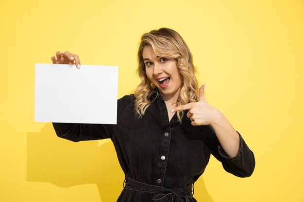 Junge schöne blonde frau im schwarzen kleid hält leeres blatt für die beschriftung auf gelbem hintergrund in den händen, die mit ihrem finger zeigen, lächelt und lacht glücklich auf gelbem hintergrund im studio