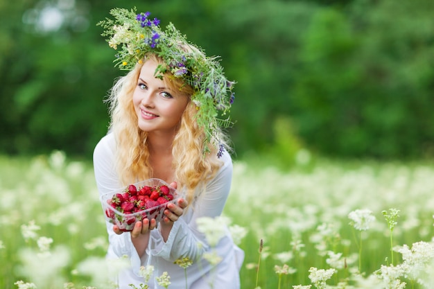 Junge schöne blonde Frau im weißen Kleid und im Blumenkranz, die Schachtel der frischen Erdbeeren am Sommertag hält