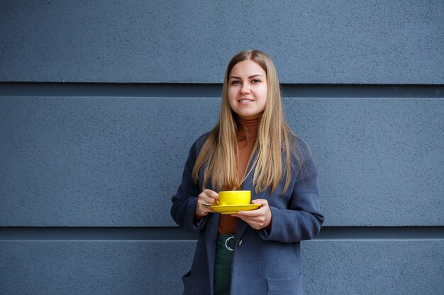 Junge schöne blonde Frau in einem grauen Mantel trinkt an einem kalten Herbsttag heißen Tee aus einer gelben Tasse. Leckeres warmes Getränk auf der Terrasse im Café