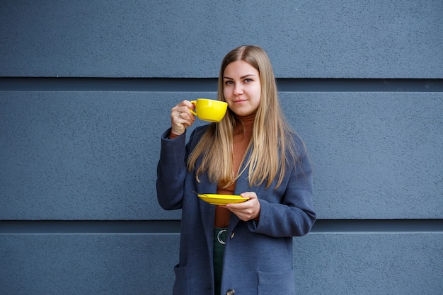 Junge schöne blonde Frau in einem grauen Mantel trinkt an einem kalten Herbsttag heißen Tee aus einer gelben Tasse. Leckeres warmes Getränk auf der Terrasse im Café